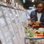 Consumidora usa o celular enquanto empurra um carrinho cheio de compras em um supermercado.