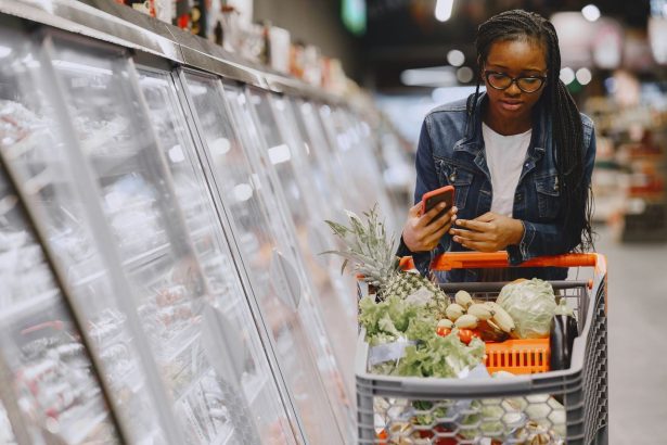 Consumidora usa o celular enquanto empurra um carrinho cheio de compras em um supermercado.