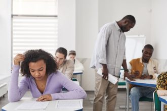 Estudantes fazem uma prova em uma sala de aula enquanto são acompanhados por uma professora.